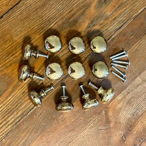 May include: A set of ten round, polished brass cabinet knobs with a smooth, reflective finish. Each knob features a central stem and a circular head. Included are ten matching silver-colored metal mounting screws arranged in a small cluster.