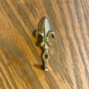 May include: A small, antique-style brass fleur-de-lis key. The key has a decorative fleur-de-lis head and a small loop at the bottom. The key is set against a wooden background.
