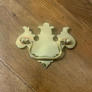 May include: A brass drawer pull with a decorative design. The pull features a central rectangular plate with a curved handle and ornate, symmetrical details on each side. The hardware is a warm gold color.