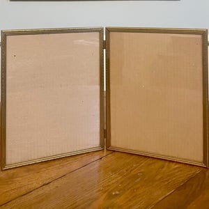 May include: A gold-toned tri-fold picture frame with two empty openings. The frame is made of metal and has a simple, classic design.