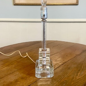 May include: A clear glass table lamp with a rectangular base and a cylindrical stem. The lamp has a silver-colored socket and a clear lightbulb. The lamp is sitting on a wooden table.