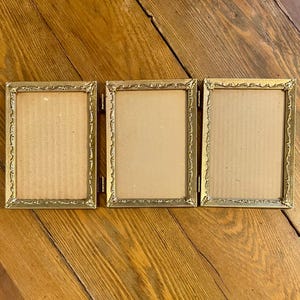 May include: A triptych picture frame with a gold-colored, ornate design. The frame is hinged, allowing it to fold. The interior of each frame is a light tan color, ready for photos or artwork. The frame is on a wooden surface.