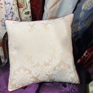 May include: A white throw pillow with a damask pattern and a red border. The pillow is sitting on a purple velvet cushion.