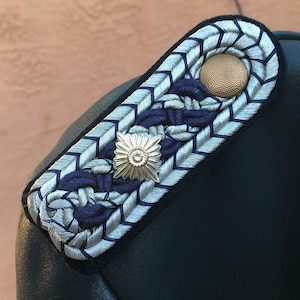 May include: Close-up of a military shoulder board with intricate silver and navy blue braiding. A gold-colored button and a starburst emblem add detail to the design. The shoulder board is set against a dark background.