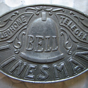 May include: Oval, antique metal sign with the words "TELEPHONE TELEGRAPH LINESMAN" around the edge. The word "BELL" is in the center, above a bell graphic. The sign is a dull silver color.