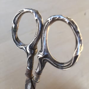 May include: Close-up of antique silver scissors with ornate, swirling handle designs. The scissors have a tarnished appearance, suggesting age and use. The word "STERLING" is visible on the handle. A vintage kitchen utensil.