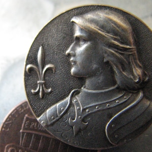May include: A silver-toned metal button featuring a profile of a knight in armor with a fleur-de-lis design. The knight is wearing a helmet and has long hair.
