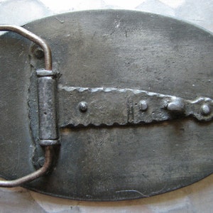 May include: A vintage metal belt buckle with an oval shape and a textured surface. The buckle features a rectangular bar with a saw-tooth edge and a metal loop for the belt. The metal has a tarnished, aged appearance.