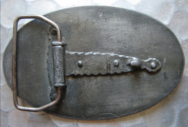 May include: A vintage metal belt buckle with an oval shape and a textured surface. The buckle features a rectangular bar with a saw-tooth edge and a metal loop for the belt. The metal has a tarnished, aged appearance.