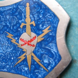 May include: A silver belt buckle with a blue enamel background. The buckle features a gold sword piercing a heart with red cross-shaped markings. The heart is surrounded by four gold lightning bolts.