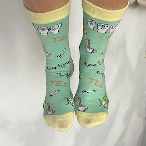 May include: A pair of light green socks with yellow cuffs and toes. The socks feature the word "TEA" in white, along with illustrations of teacups, spoons, and tea leaves. The phrase "tea time" is also printed on the socks.