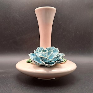 May include: A pink ceramic vase with a light blue flower accent. The vase has a wide, rounded base and a tall, slender neck. The flower is a detailed, three-dimensional design with green leaves.