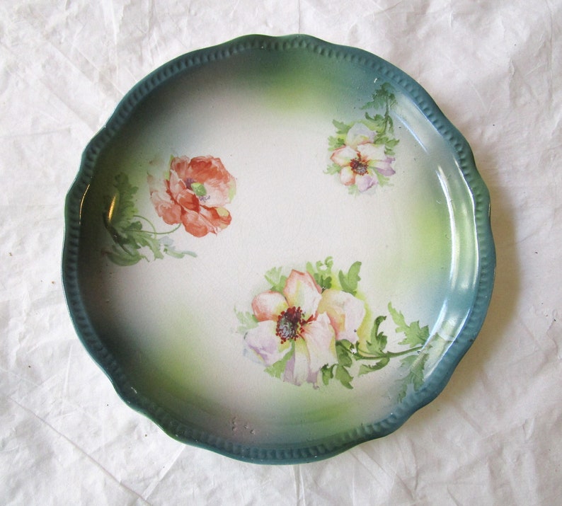May include: A vintage ceramic plate with a scalloped edge and a green glaze. The plate features a hand-painted floral design with pink, white, and red flowers.