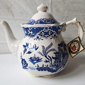 Puede incluir: Una tetera de cerámica blanca con un patrón azul de estilo willow que muestra pájaros, un barco y elementos arquitectónicos. La tapa tiene un borde azul a juego. Una etiqueta unida al asa dice Price and Kensington Made in England.