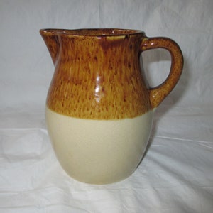 Puede incluir: Una jarra de cerámica esmaltada en marrón y crema con cuerpo redondeado y asa. La jarra tiene un esmalte marrón en la mitad superior y un esmalte crema en la mitad inferior.
