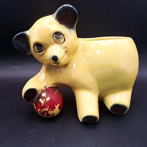 May include: A yellow ceramic bear planter with a glossy finish, featuring dark brown accents on the ears and paws. The bear is depicted in a standing position, resting one front paw on a round, textured red ball with white distressed markings.
