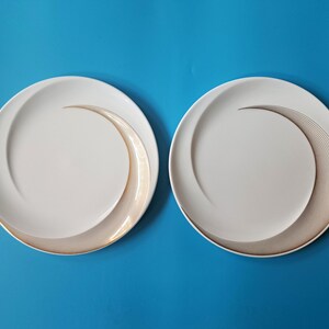 May include: Two round white ceramic plates featuring a smooth, curved indentation on one side. Each plate is decorated with a series of fine, concentric bronze-colored lines that follow the curve of the indentation, creating a swirling pattern.