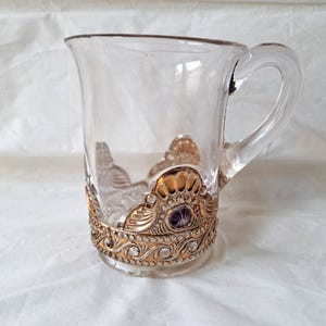 Puede incluir: Una taza de cerámica blanca con un asa negra y el texto "But First, Coffee" en negro, en negrita. La taza está sobre una superficie blanca.