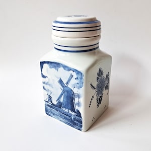 Puede incluir: Un recipiente de cerámica cuadrado con tapa extraíble, que presenta una base blanca con ilustraciones pintadas a mano en azul. Un lado muestra un paisaje de molino de viento, mientras que el otro lado muestra un motivo floral.