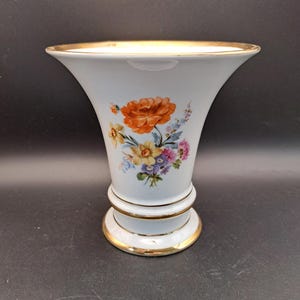 Może przedstawiać: Biały ceramiczny wazon z rozkloszowaną górą i złotą obwódką. Wazon posiada kwiatowy wzór z pomarańczowymi, żółtymi, różowymi i niebieskimi kwiatami. Podstawa i środkowa część wazonu są również ozdobione złotymi obramowaniami.