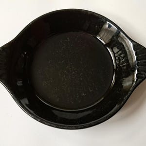 May include: A black, round ceramic dish with two handles. The dish has a glossy finish and a slightly textured surface. The handles are curved and have a ridged design. The dish is set against a white background.