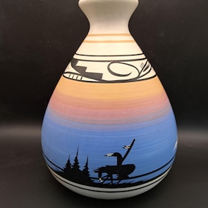 Peut inclure: Un vase en céramique peint à la main, avec une base large et un col étroit. Le vase présente un dégradé de couleurs, passant du bleu à l'orange. Une silhouette noire d'oiseau et d'arbres décore la section bleue. Le haut est blanc avec des motifs noirs et beiges.