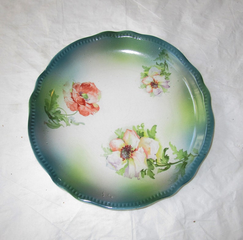 May include: A vintage porcelain plate with a scalloped edge and a green and white glaze. The plate features a hand-painted floral design with pink, white, and green flowers.