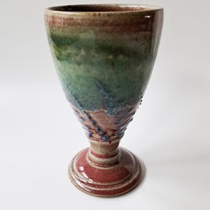 Puede incluir: Una copa de cerámica hecha a mano con una parte superior acampanada y una base con pie. La copa presenta una mezcla de colores, incluyendo verde, azul y rojo, con un acabado texturizado y vidriado. La base es de color rojo intenso con anillos concéntricos.