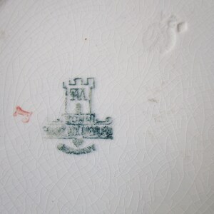 May include: A close-up of a white ceramic plate with a green and white mark. The mark features a castle with the letters "RM" inside and the words "Royal Doulton" below.