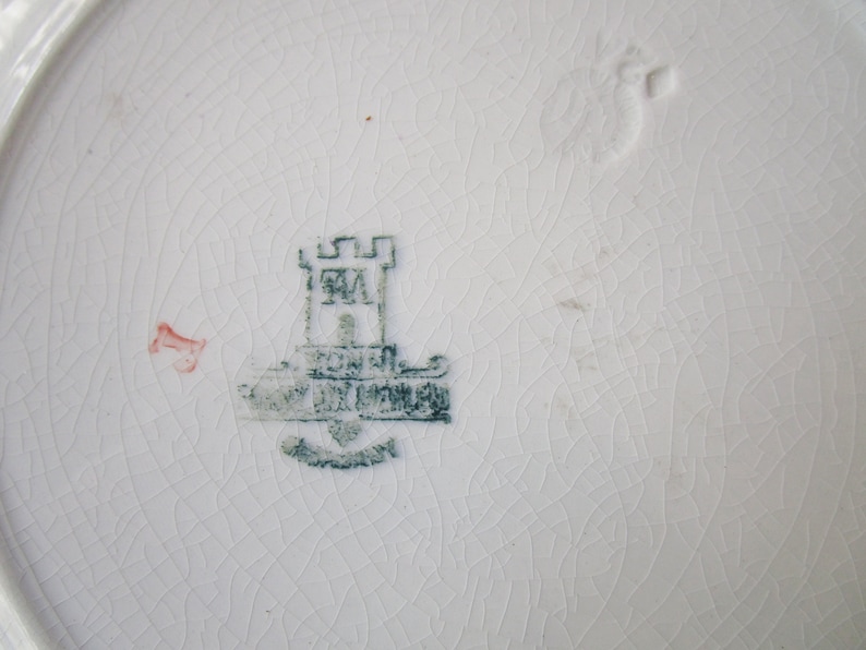 May include: A close-up of a white ceramic plate with a green and white mark. The mark features a castle with the letters "RM" inside and the words "Royal Doulton" below.