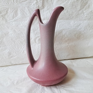 May include: A pink ceramic jug with a distinctive, curved handle. The jug has a speckled glaze and a smooth, rounded base.