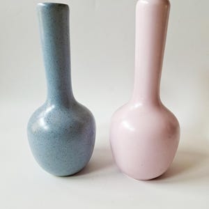 May include: Two ceramic bud vases with narrow necks and rounded bases. One vase is a speckled light blue color, while the other vase is a solid, smooth light pink color. Both items feature a simple, vertical design with a small opening at the top.