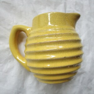 Puede incluir: Jarra de cerámica amarilla con diseño acanalado y asa. La jarra tiene un pico para verter y un acabado craquelado. La jarra es de color amarillo brillante y probablemente se utiliza para servir bebidas.