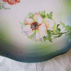 May include: A vintage ceramic plate with a scalloped edge and a green and white glaze. The plate features a hand-painted floral design with pink and white flowers and green leaves.