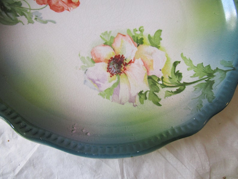 May include: A vintage ceramic plate with a scalloped edge and a green and white glaze. The plate features a hand-painted floral design with pink and white flowers and green leaves.