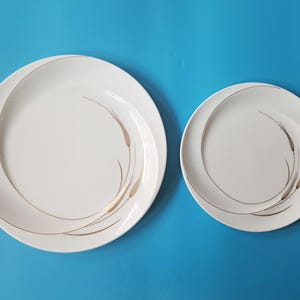 Könnte beinhalten: Zwei weiße, runde Keramikteller in verschiedenen Größen, die jeweils mit einem geschwungenen, goldenen Liniendesign verziert sind, das sich in einer kreisförmigen Bewegung über die Oberfläche zieht und eine elegante Optik bietet.