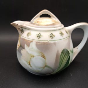 May include: Cream-colored ceramic creamer with a lid, featuring a floral design with white flowers and green leaves. The creamer has a curved handle and a spout for pouring. The lid has a small opening for steam release.