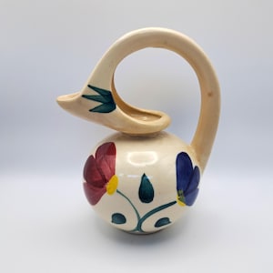 Könnte beinhalten: Cremefarbener Keramikkrug mit kugelförmigem Boden und gebogenem Henkel. Der Krug ist mit roten, blauen und gelben Blumendesigns und grünen Blättern verziert. Der Ausguss ist wie eine kleine Rinne geformt.