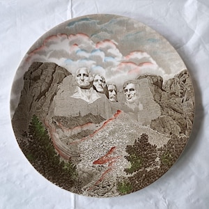 Puede incluir: Un plato decorativo redondo con una imagen del Monte Rushmore. El plato muestra los rostros de cuatro presidentes estadounidenses tallados en una montaña, con un paisaje detallado y un cielo nublado. El plato tiene un esquema de color marrón y gris.