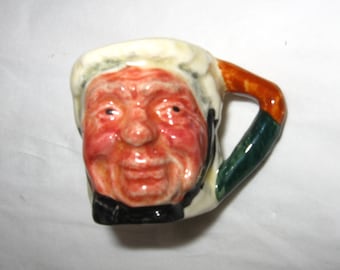 May include: A small ceramic mug with a face of an older person. The mug has a green and brown handle and is decorated with white and yellow paint.