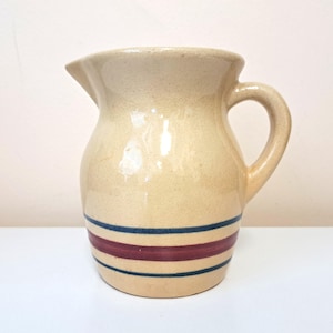 Puede incluir: Una jarra de cerámica de color crema con un diseño de rayas azules y burdeos. La jarra tiene un cuerpo redondeado y un asa.