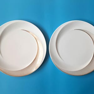 Op de afbeelding: Twee ronde witte keramische borden met een glad, concaaf midden en een brede, gebogen rand versierd met een reeks fijne, concentrische gouden lijnen die een wervelend, halvemaanvormig patroon vormen aan de rechterkant.