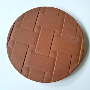 May include: A round, terracotta-colored coaster with a woven basket pattern. The coaster is made of a solid material and has a slightly raised texture. The design is a repeating pattern of rectangular shapes.