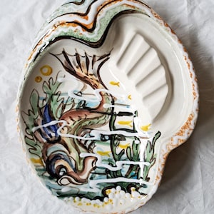Könnte beinhalten: Eine keramische, muschelförmige Schale mit einer handgemalten Unterwasserszene. Die Schale zeigt einen Fisch, Seetang und kleine gelbe Fische. Der Rand hat ein braunes und orangefarbenes Muster. Die Schale ist etwa 25 cm lang.