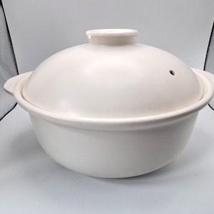 Könnte beinhalten: Ein weißer Keramikkochtopf mit passendem Deckel. Der Topf hat zwei Griffe und eine abgerundete Form. Der Deckel hat einen kleinen Knopf und ein Entlüftungsloch. Der Topf ist zum Kochen und Servieren bestimmt.