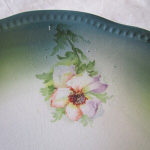 May include: A vintage ceramic plate with a green rim and a white center featuring a hand-painted floral design. The flower is a light pink and purple with a red center.