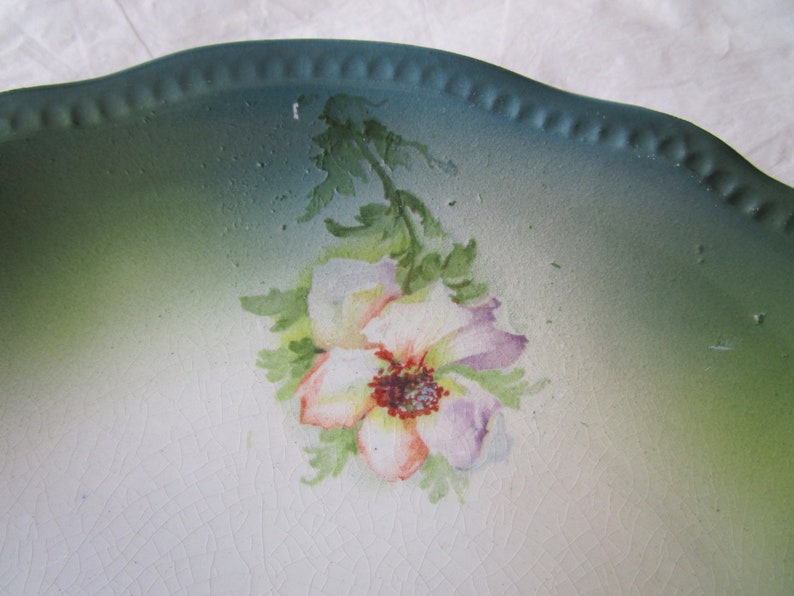 May include: A vintage ceramic plate with a green rim and a white center featuring a hand-painted floral design. The flower is a light pink and purple with a red center.