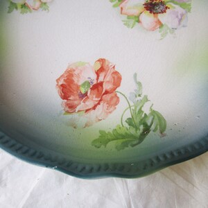 May include: A vintage ceramic plate with a scalloped edge and a green and white glaze. The plate features a hand-painted floral design with red, pink, and white flowers.
