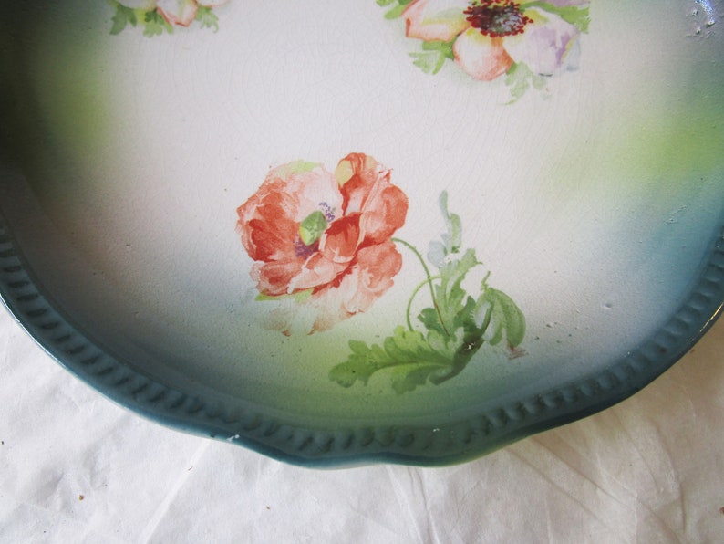 May include: A vintage ceramic plate with a scalloped edge and a green and white glaze. The plate features a hand-painted floral design with red, pink, and white flowers.