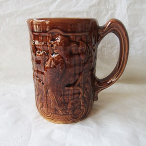 Pode incluir: Uma caneca de cerâmica marrom com um design em relevo de pássaros e galhos. A caneca tem uma alça grande.